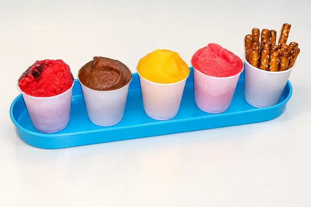 Five cups of Italian ice in a blue tray with pretzel sticks